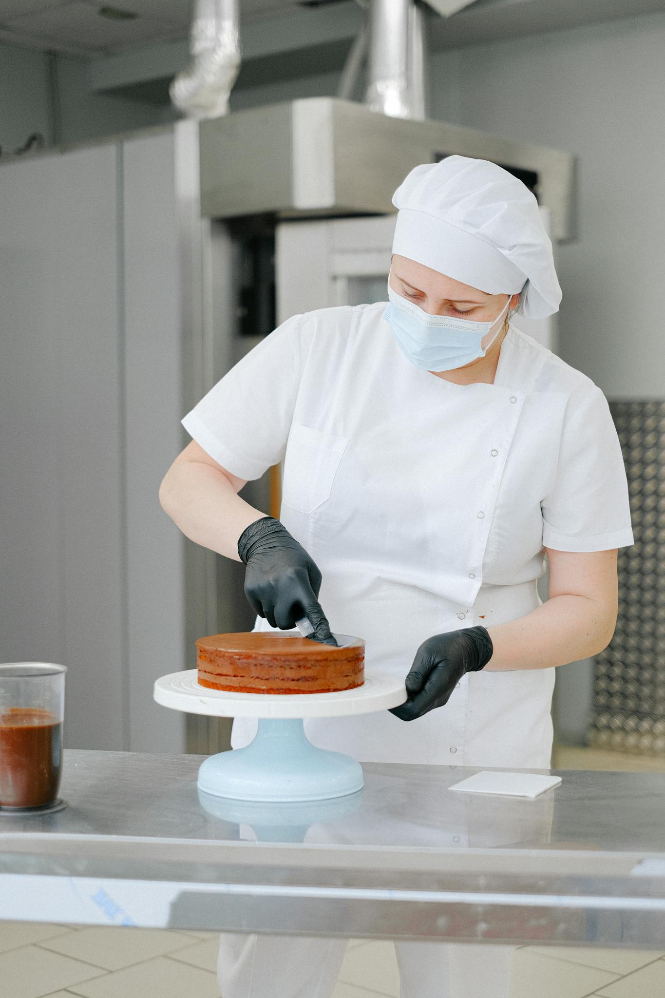 A skilled baker in a white uniform decorates a chocolate cake in a commercial kitchen.