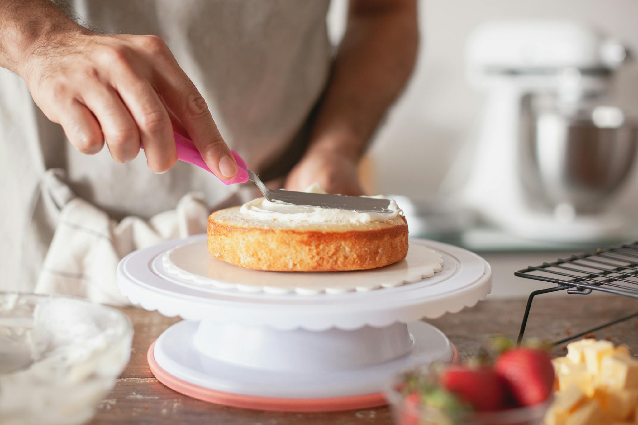 Close-up of person decorating a cake with frosting using a pink spatula on a cake stand.