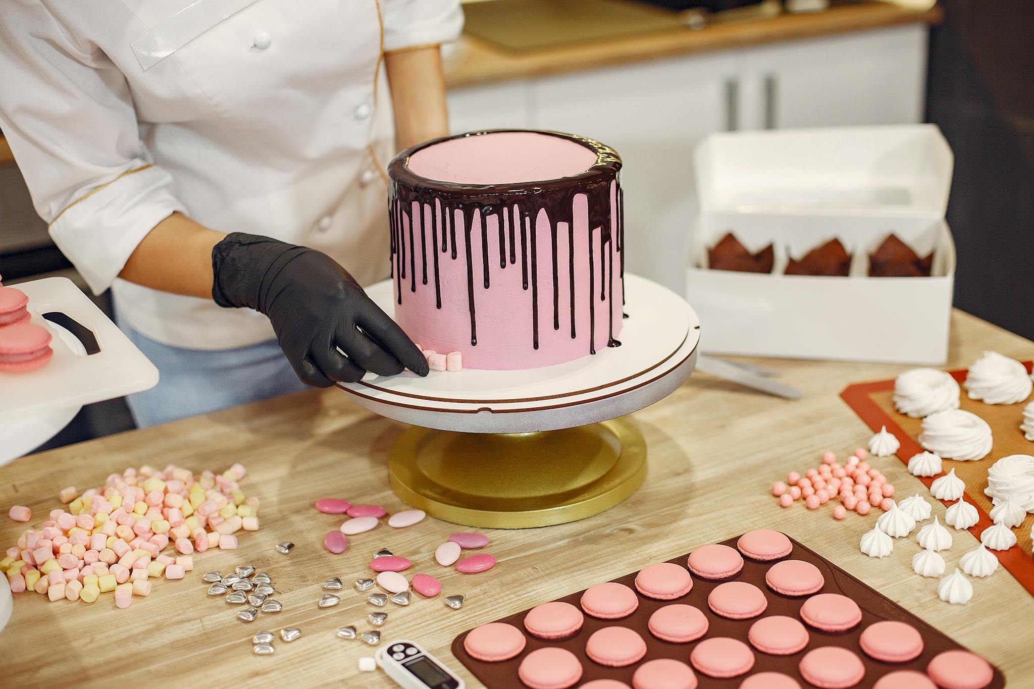 From above crop female cook in uniform and latex gloves placing small sweets on bottom of cake placed on stand near desserts in confectionery