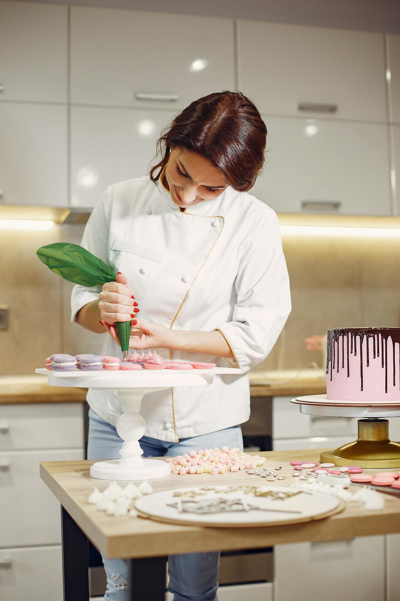 Positive female pastry cook in white apron pouring cream on yummy macaroons placed on white stand while working in modern laboratory